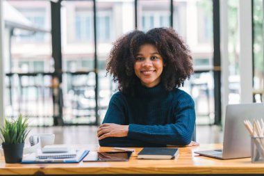 Mutlu bir Afro-Amerikan kadının portresi, özgüvenle gülümseyen, afro saçlı, oturan ve modern bir ofiste çalışan