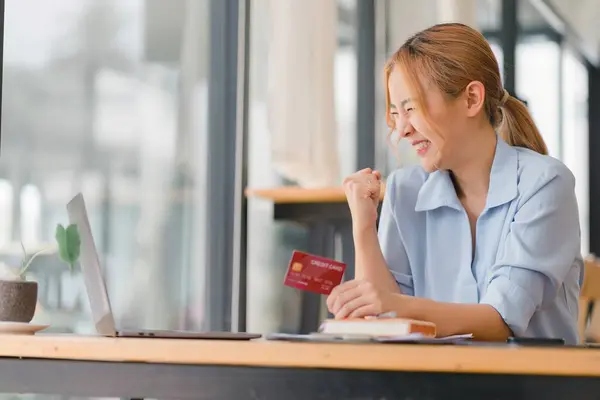Smiling millennial woman holds a credit card, engaged in online laptop shopping at a cafe. A happy female shopper purchases goods or services in an internet store