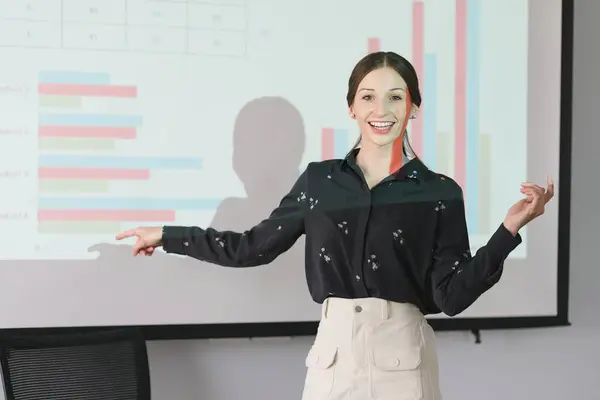 The female Chief Analyst is conducting a meeting presentation for a team of economists. The projector screen shows graphs, product sales, revenue growth strategies, and e-commerce analysis