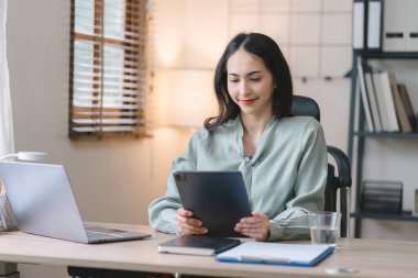 Modern bir ofisteki satış verilerini analiz etmek için dijital tablet kullanan gülümseyen, genç, güzel, profesyonel ve kendinden emin Asyalı bir iş kadınının portresi.