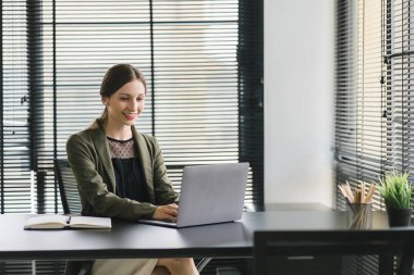 Şık elbiseli başarılı bir iş kadını modern bir ofiste masa başında dizüstü bilgisayar kullanıyor. İş projelerini planlayan başarılı bir finans yöneticisi gibi görünüyor.