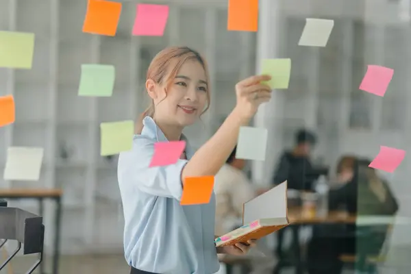 A millennial Asian businesswoman shares ideas using sticky notes, which she places on a glass wall