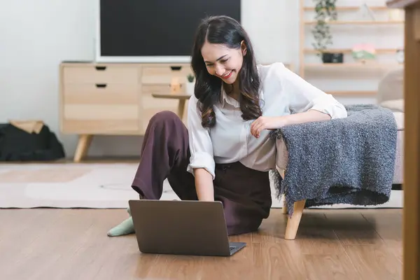 An attractive millennial businesswoman is sitting on the floor in her living room, working with a laptop computer at home