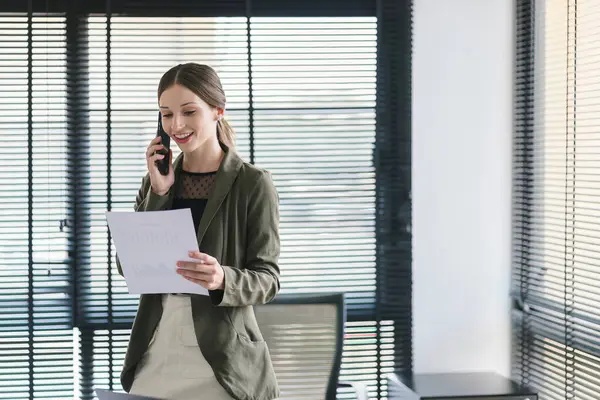 Happy professional millennial businesswoman in casual attire is making a business call while analyzing financial data on financial report at her office