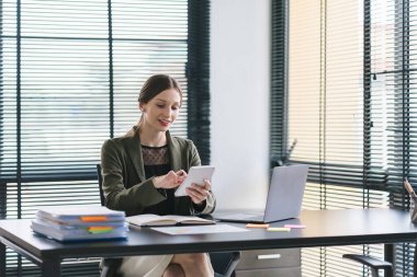 Modern bir ofisteki satış verilerini analiz etmek için dijital tablet kullanan gülümseyen, genç, güzel, profesyonel ve kendinden emin bir beyaz iş kadınının portresi.