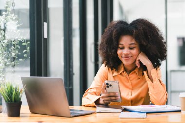 Bir dizüstü bilgisayar üzerinde çalışırken akıllı telefon kullanan Afrikalı bir iş kadını. Gülümseyen Afrikalı Amerikalı kadın oyun oynamak için cep telefonu uygulaması kullanıyor, online alışveriş yapıyor, ofiste.