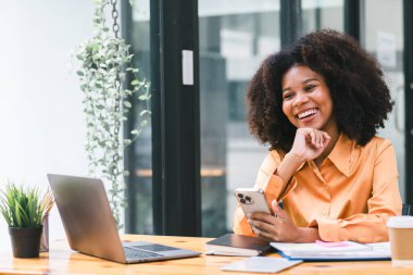 Bir dizüstü bilgisayar üzerinde çalışırken akıllı telefon kullanan Afrikalı bir iş kadını. Gülümseyen Afrikalı Amerikalı kadın oyun oynamak için cep telefonu uygulaması kullanıyor, online alışveriş yapıyor, ofiste.