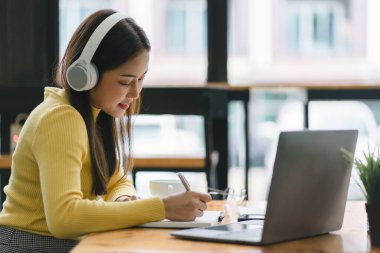 Kulaklık takan Asyalı iş kadını Webinar video kursunu izlerken not alıyor. İnternette okumak için e-öğrenim yoluyla ders dinleyen ciddi bir kız öğrenci.