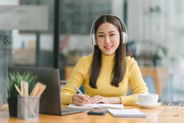 Kulaklık takan Asyalı iş kadını Webinar video kursunu izlerken not alıyor. İnternette okumak için e-öğrenim yoluyla ders dinleyen ciddi bir kız öğrenci.