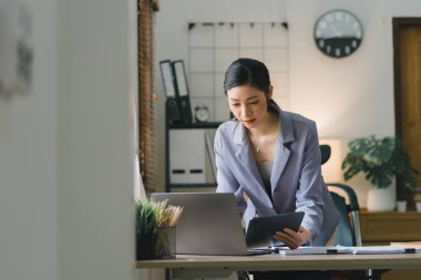 Modern bir ofisteki satış verilerini analiz etmek için dijital tablet kullanan gülümseyen, genç, güzel, profesyonel ve kendinden emin Asyalı bir iş kadınının portresi.