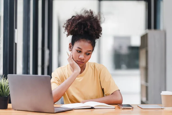 Young African American woman is taking notes in a notebook while watching a webinar video course. Afro american student listening to the lecture to study online through e-learning