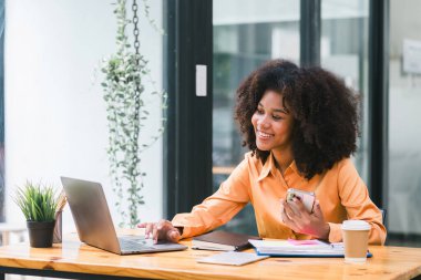 Neşeli, Afro-Amerikan güzel bir kadın, gündelik giyinen, gülümseyen, akıllı telefon kullanan ve modern bir ofiste çalışan bir girişimci.