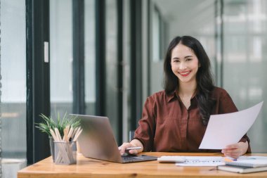 Asyalı bir iş kadını dizüstü bilgisayar kullanıyor ve hesap makinesi ve belgelerle çalışıyor, yatırımlar için mali planlama yapıyor ve mali raporu analiz ediyor