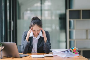 Stresli ve yorgun Asyalı iş kadını ofis masasında eli başının üstünde otururken görülüyor. Bu da yoğun bir iş günü geçirdiğini gösteriyor.