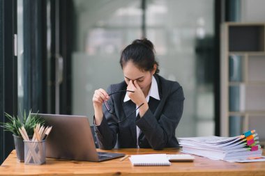 Stresli ve yorgun Asyalı iş kadını ofis masasında eli başının üstünde otururken görülüyor. Bu da yoğun bir iş günü geçirdiğini gösteriyor.