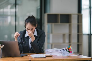 Stresli ve yorgun Asyalı iş kadını ofis masasında eli başının üstünde otururken görülüyor. Bu da yoğun bir iş günü geçirdiğini gösteriyor.