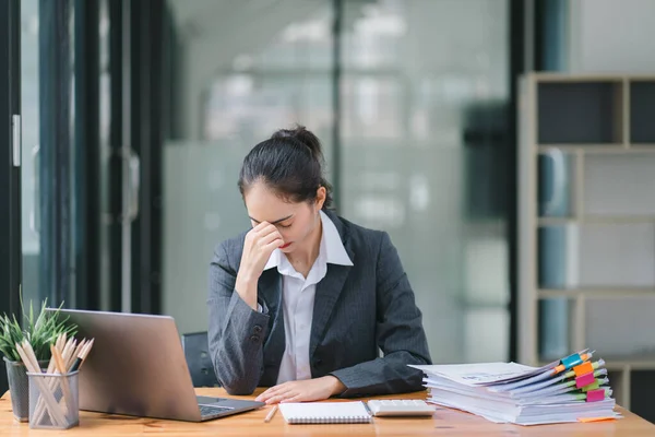 The stressed and exhausted millennial Asian businesswoman is seen sitting at her office desk with her hand on her head, indicating a hard working day where she is overloaded with work