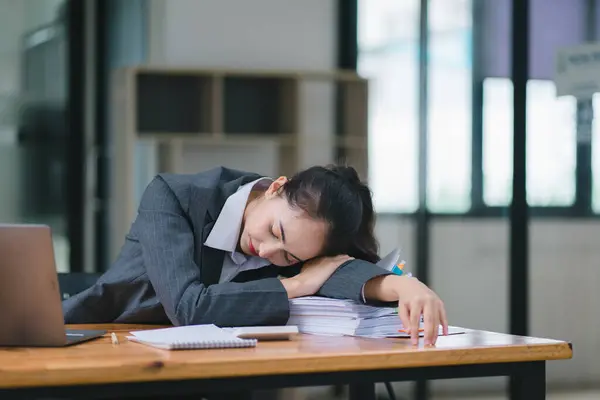 The tired millennial Asian businesswoman is feeling sleepy and bored from sitting at her desk for a long time, indicating a hard working day where she is overloaded with work
