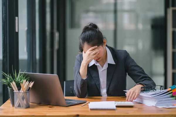 The stressed and exhausted millennial Asian businesswoman is seen sitting at her office desk with her hand on her head, indicating a hard working day where she is overloaded with work