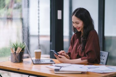 Melez, Asyalı bir iş kadını modern bir ofiste masasında akıllı telefon kullanıyor.