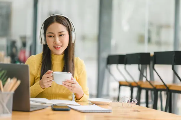 Asian businesswoman wearing headphones is taking notes in a notebook while watching a webinar video course. Happy female student listening to the lecture to study online through e-learning
