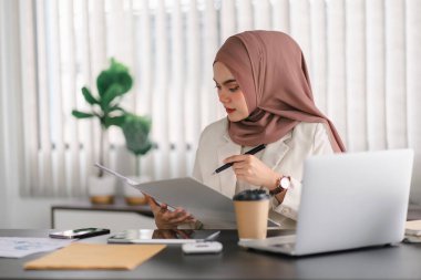 Kahverengi başörtülü Asyalı Müslüman kadın modern ofiste dizüstü bilgisayarla çalışıyor.