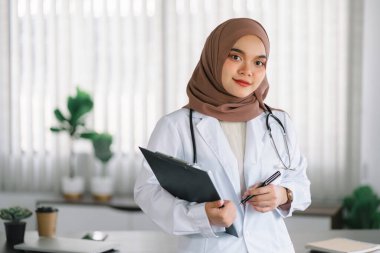 Kahverengi tesettürlü gülümseyen Müslüman kadın doktor tıbbi kayıtlar taşıyor.