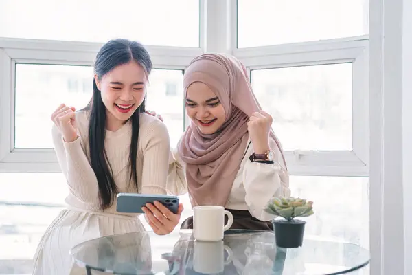 Two cheerful young Asian women raise arms in celebration together while looking at a smartphone screen