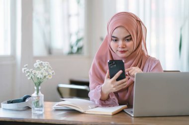 Ofiste bilgisayar başında çalışırken tesettürlü Asyalı Müslüman kadın telefon kullanıyor.