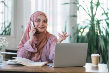 Ofiste bilgisayar başında çalışırken tesettürlü Asyalı Müslüman kadın telefon kullanıyor.