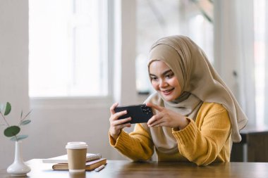 Asyalı Müslüman bir iş kadını kafeteryada dinlenirken telefon kullanarak tesettüre giriyor.