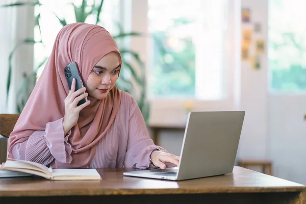 Ofiste bilgisayar başında çalışırken tesettürlü Asyalı Müslüman kadın telefon kullanıyor.