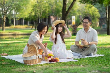 Mutlu bir aile parkta piknik yapıyor. Aileler ve çocuklar çimlerin üzerinde oturuyor ve güneşli bir yaz gününde dışarıda sağlıklı yemeklerin tadını çıkarıyor.