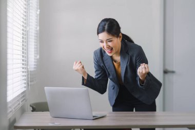 Triumphant Asian businesswoman celebrating professional victory with arms raised in front of a laptop at home office