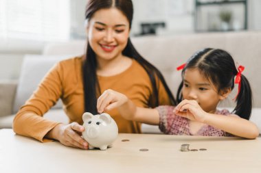 Küçük tatlı Asyalı kız para biriktirmek için annesiyle birlikte kumbaraya para koyuyor..