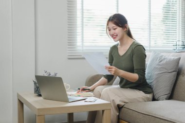 Mutlu Asyalı genç iş kadını vergileri hesaplıyor, yerli faturaları sayıyor, e-bankacılık, e-ticaret, evde hesaplama için dizüstü bilgisayar kullanıyor..
