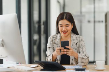 Gülümseyen iş kadını modern ofis masasında akıllı telefonu kontrol ediyor.