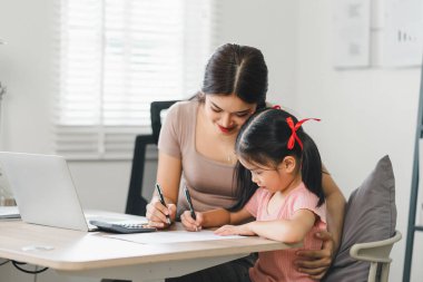 Anne, kızının ödevlerini yapmasına yardım ediyor. Masada oturup kağıt deftere doğru cevabı yazıyor.