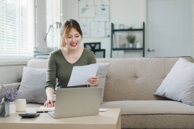 Mutlu Asyalı genç iş kadını vergileri hesaplıyor, yerli faturaları sayıyor, e-bankacılık, e-ticaret, evde hesaplama için dizüstü bilgisayar kullanıyor..