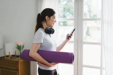 Neşeli bir kadın oturma odasında elinde mor yoga minderiyle ve kulaklık takarak yoga seansına hazırlanıyor..