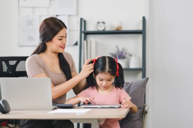 Anne, kızına dijital tabletle yardım ederken evde çalışıyor, iş ve aile zamanını modern bir ev ofisinde dengeliyor.