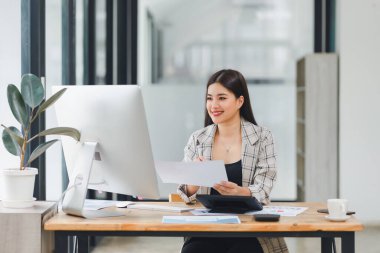 Neşeli Asyalı iş kadını elinde belgelerle modern ofisteki bilgisayarda çalışıyor.