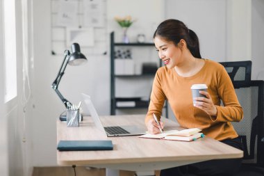 Evde dizüstü bilgisayar ve kahveyle çalışan gülümseyen kadın notlar alıyor ve verimli çalışma alanlarının tadını çıkarıyor.
