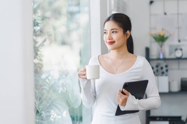 Tablet ve kahve fincanı tutan, gülümseyen kadın, pencerenin yanında duran, dışarı bakan, aydınlık, modern bir ev