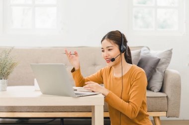 Kulaklık takan gülümseyen kadın, evde bilgisayarla çalışırken video çağrısında bulundu ve notlar aldı.