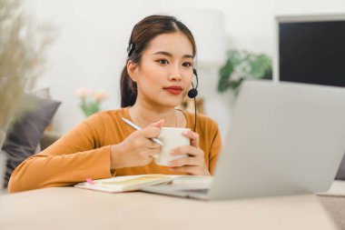 Kulaklık takan bir kadın, aktif olarak çevrimiçi bir öğrenme oturumunda bilgisayar ekranında gösterilen bir öğretmenle evden ders çalışıyor..