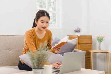 Dizüstü bilgisayarda çalışan gülümseyen kadın, evinde defter ve belgelerle oturuyor.