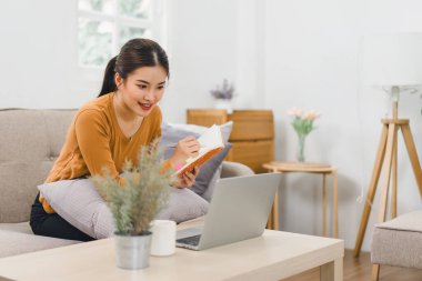Dizüstü bilgisayarda çalışan gülümseyen kadın, evinde defter ve belgelerle oturuyor.
