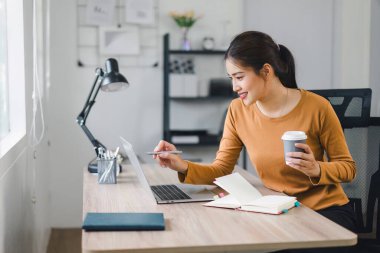 Dizüstü bilgisayarda çalışan gülümseyen kadın defter ve belgelerle rahat bir ofiste otururken....