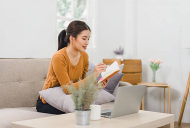 Dizüstü bilgisayarda çalışan gülümseyen kadın, evinde defter ve belgelerle oturuyor.